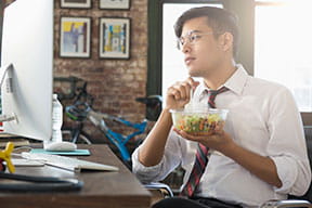 employee holding his lunch in a clear bowl, looking thoughtfully at his computer monitor