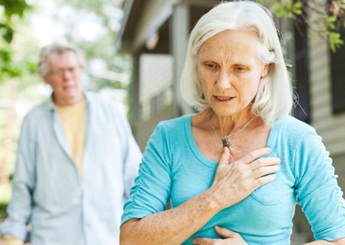 older woman looking concerned, holding her hand over her chest, while older man approaches behind her