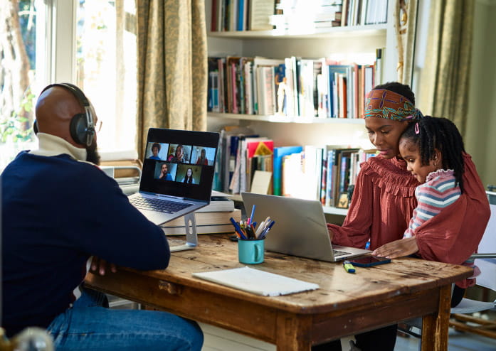 black mom and dad sitting at table in house looking at laptops. mom is holding toddler daughter in her lap.